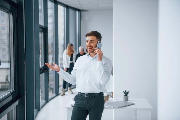 Homem em roupas formais falando por telefone e na frente de uma jovem equipe de sucesso que trabalha e se comunica juntos dentro de casa no escritório