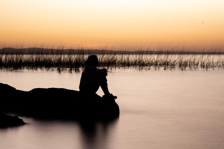 Foto homem em silhueta sentado ao lado do lago contra o céu durante o pôr do sol