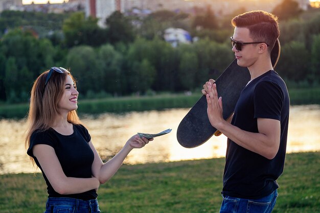 Homem em sunflasses segura um skate (longboard) e a mulher oferece dinheiro ao pôr do sol na praia. compra e venda de skate esportivo no parque de verão