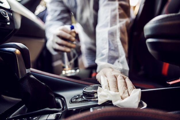 Homem em traje de proteção com máscara desinfetante dentro do carro, limpe as superfícies que são tocadas com frequência, evite infecção de coronavírus, contaminação de germes ou bactérias. Infecção