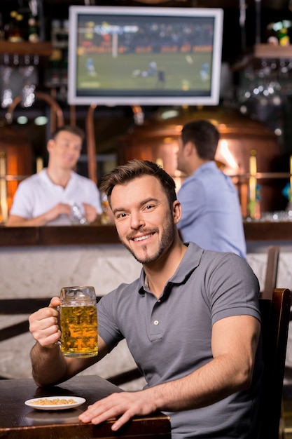 Homem em um bar de cerveja. Jovem bonito segurando uma caneca de cerveja e sorrindo enquanto está sentado em um bar