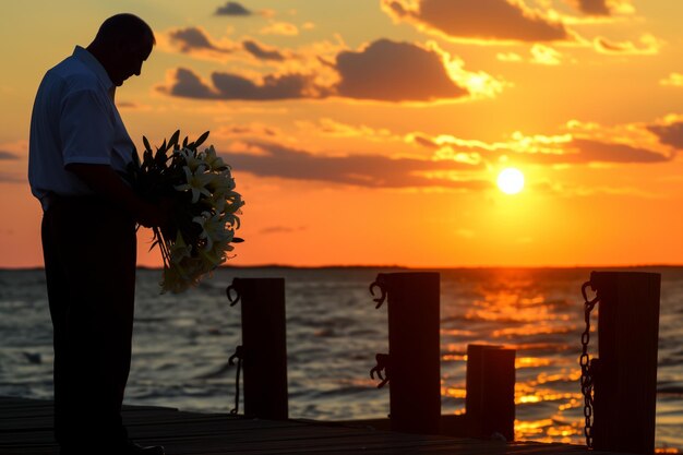 Foto homem em um cais ao pôr-do-sol segurando um bando de lírios