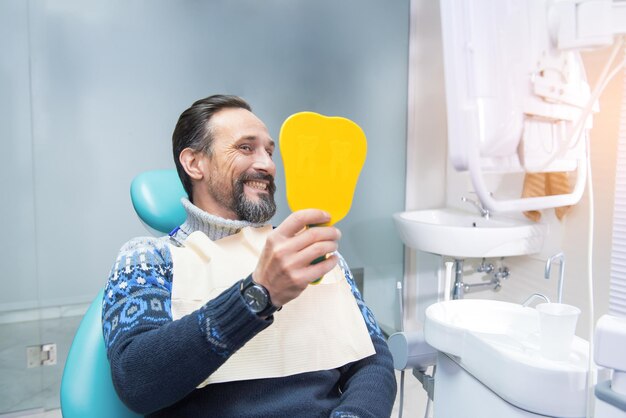 Homem em uma cadeira odontológica sorrindo e segurando o espelho torna seu sorriso perfeito