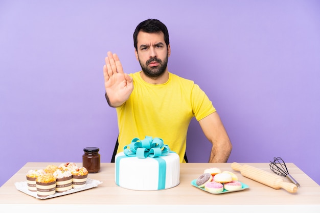 Homem em uma mesa com um bolo grande, fazendo o gesto de parada