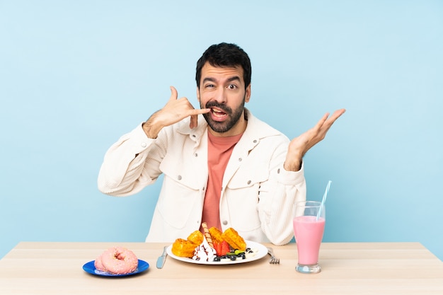 Homem em uma mesa tomando waffles de café da manhã e um milk-shake fazendo gesto de telefone e duvidando