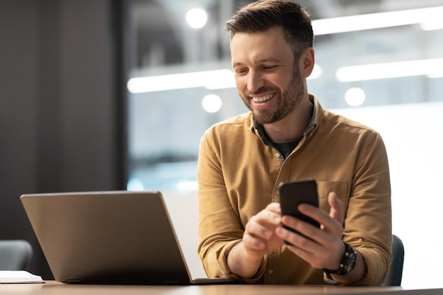 Homem empresário feliz enviando mensagens de texto usando smartphone perto de laptop no escritório