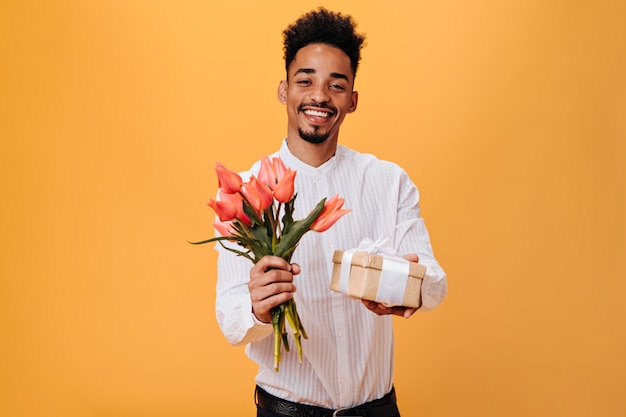 Homem encantador posa alegremente na parede laranja e segurando flores, presente