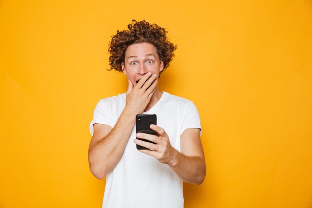 Homem encaracolado chocado ou surpreso em casual camiseta branca gritando enquanto segura o smartphone