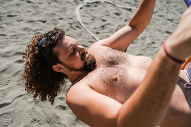 Homem encaracolado e barbudo malhando na barra horizontal na praia