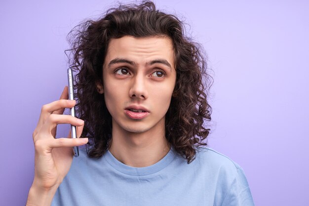 Homem encaracolado incerto falando em smartphone, ouvindo falar, cara jovem com cara séria, em camiseta casual, isolado no fundo roxo