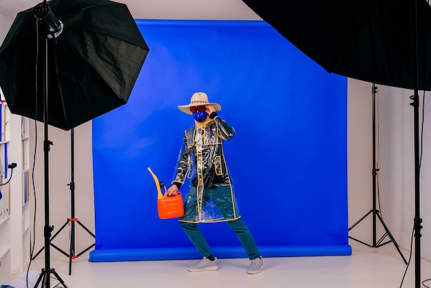 Homem engraçado elegante e estranho com máscara e chapéu de palha com regador vermelho posando sobre uma parede azul