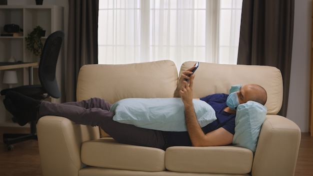 Homem entediado com máscara de proteção deitado no sofá navegando no smartphone durante o auto-isolamento do coronavírus.