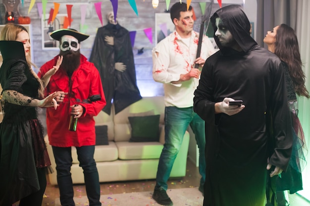 Homem entediado vestido como um ceifador na festa de halloween usando seu telefone.