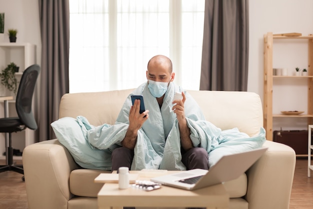 Homem envolto em um cobertor, segurando o frasco de comprimidos em uma videochamada com um médico durante a pandemia global.