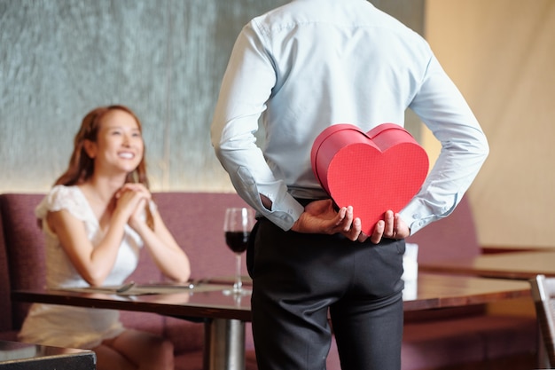 Foto homem escondendo um presente romântico para a namorada nas costas quando está à mesa do restaurante