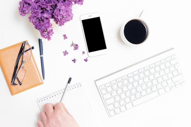 Homem escrevendo notas no diário. Local de trabalho com teclado de computador, telefone celular, xícara de café, caderno, caneta, lápis, óculos e flores lilás no fundo da mesa branca. Postura plana, vista superior