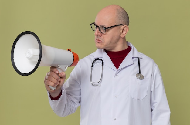 Homem eslavo adulto impressionado com óculos ópticos em uniforme de médico com estetoscópio segurando e olhando para o alto-falante