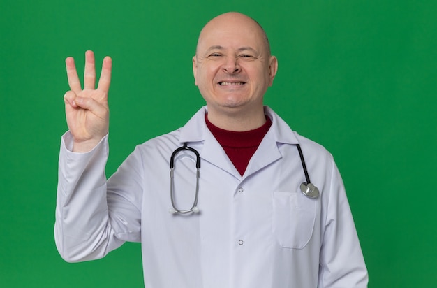 Homem eslavo adulto sorridente com uniforme de médico e estetoscópio gesticulando três com os dedos