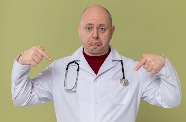 Homem eslavo adulto triste com uniforme de médico e estetoscópio apontando para baixo