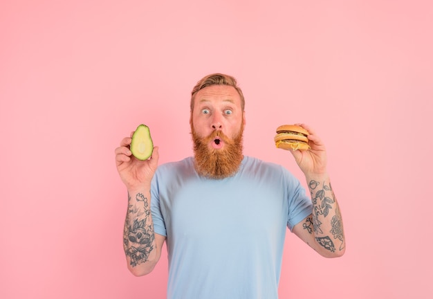 Homem espantado com barba e tatuagens está indeciso se comer um abacate ou um hambúrguer