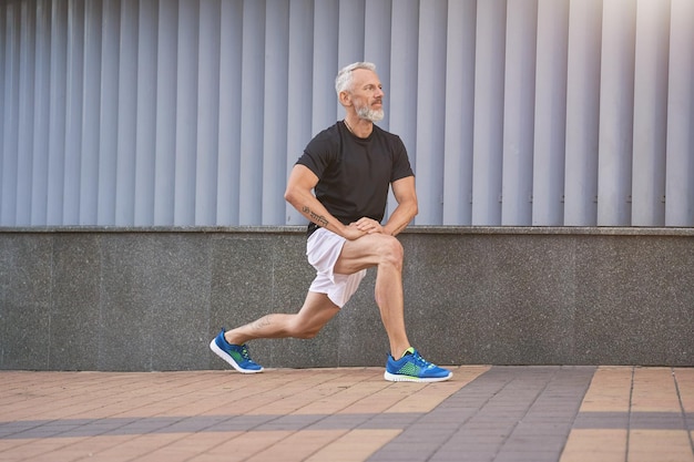 Homem esportivo de meia-idade ativo em roupas esportivas, esticando as pernas, aquecendo e se exercitando