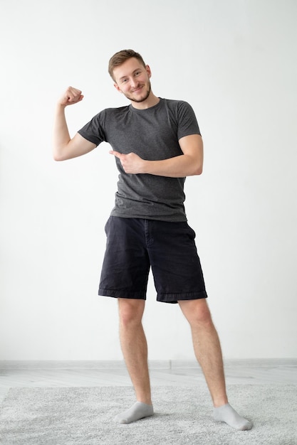 Homem esportivo feliz Corpo forte Fitness em casa Resultados da academia Cara autoconfiante em shorts de camiseta mostrando os músculos do bíceps mãos no fundo interior da parede clara