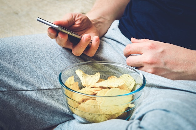 homem está descansando no smartphone sofwith comendo batatas fritas de prato de vidro.