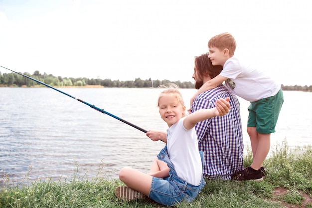 Homem está sentado na grama e segurando a vara de peixe. Ele está tentando pegar algum peixe. Rapaz está de pé atrás e abraçando-o. Menina está olhando para trás e acenando.