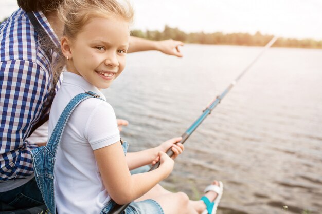 Homem está sentado na margem do rio com sua filha e apontando para a frente. Menina está segurando a vara de peixe e olhando para a câmera. Ela está sorrindo. Menina parece feliz.