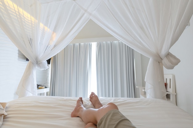Homem esticando as pernas com mosquiteiro na cama nas férias, luz do sol através da cortina branca no quarto