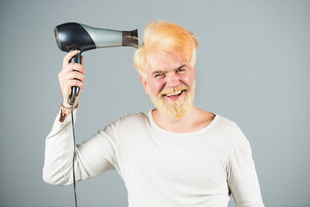 Homem estiloso com secador de cabelo e expressões engraçadas na barbearia. Cabelo loiro barbudo seco.