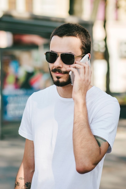 Homem estiloso com tatuagem e barba, usando óculos e uma camiseta branca, usa seu smartphone