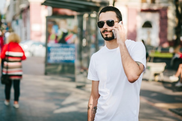 Homem estiloso com tatuagem e barba, usando óculos e uma camiseta branca, usa seu smartphone