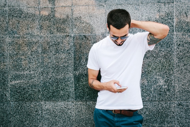 Homem estiloso com tatuagem e barba, usando óculos e uma camiseta branca, usa seu smartphone