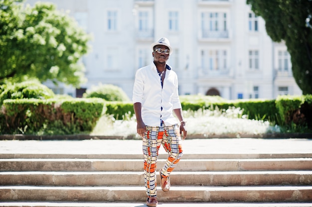 Homem estiloso na camisa branca e calça colorida com chapéu e óculos colocados ao ar livre