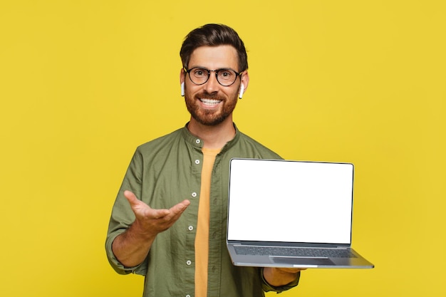 Homem estiloso positivo mostrando um novo laptop com tela branca em branco sobre a maquete de espaço de cópia de fundo de estúdio amarelo