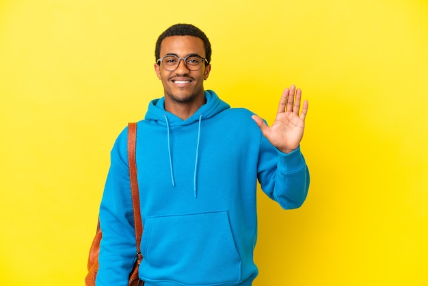 Homem estudante afro-americano sobre fundo amarelo isolado saudando com a mão com expressão feliz