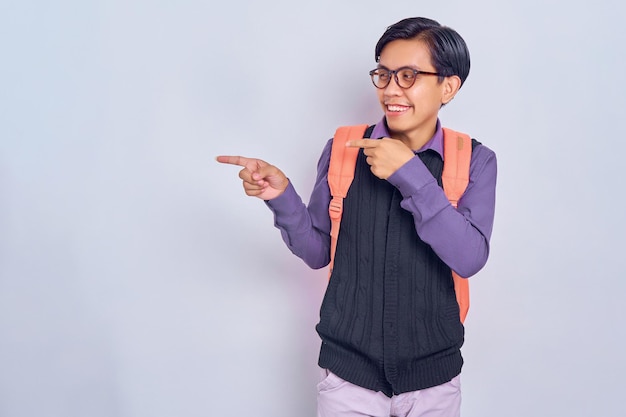 Homem estudante asiático positivo apontando o dedo para longe com um sorriso cheio de dentes no rosto espaço vazio para o seu anúncio isolado em fundo cinza