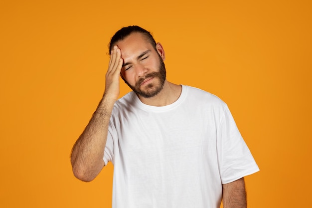 Homem europeu adulto chateado com barba em camiseta branca pressiona mão na cabeça sofre de enxaqueca