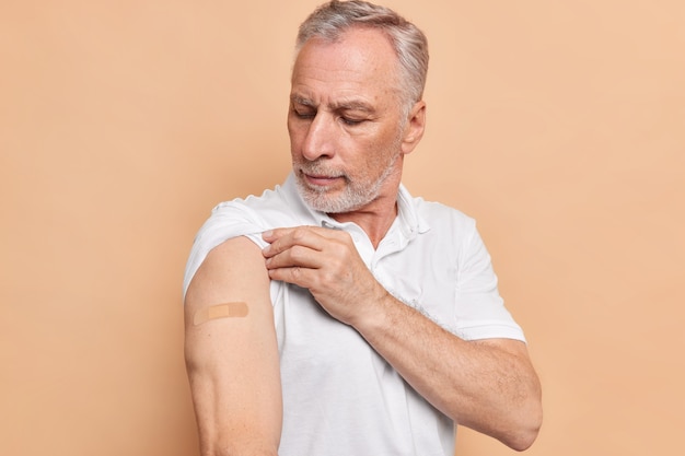 Homem europeu barbudo olha para o braço com gesso satisfeito com vacinas contra o coronavírus, que é seguro e eficaz, usa camiseta branca isolada na parede bege