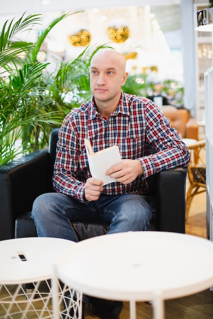 Homem europeu careca com uma camisa xadrez vermelha e jeans azul sentado em uma poltrona em um apartamento bem iluminado com muitas plantas verdes, lendo um livro em um ambiente caseiro relaxante