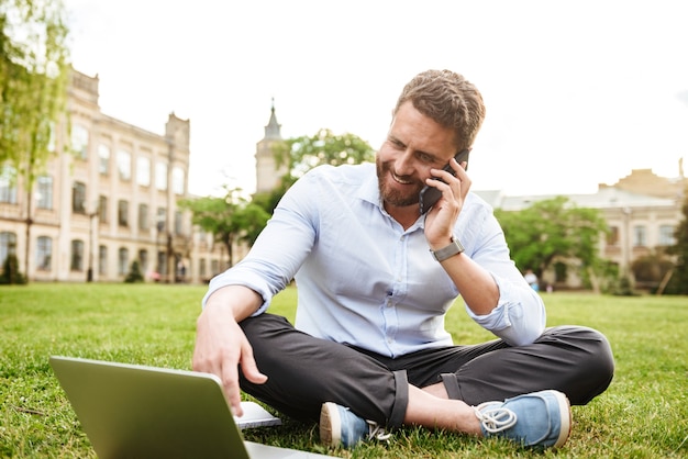 homem europeu de conteúdo em roupas de negócios, sentado na grama do parque com as pernas cruzadas e falando no celular enquanto usa um laptop prateado