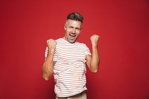 Homem europeu em camiseta listrada gritando e cerrando os punhos isolados no vermelho