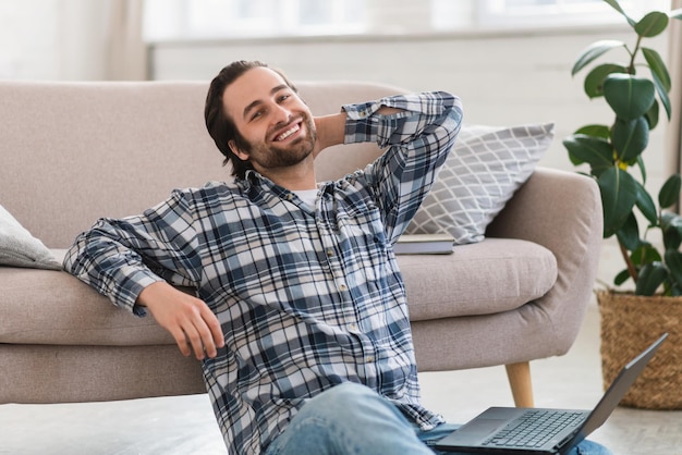 Homem europeu milenar feliz em roupas domésticas com barba por fazer senta-se no chão trabalhando no computador na sala de estar