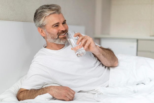 Foto homem europeu positivo sentado na cama segurando um copo de água