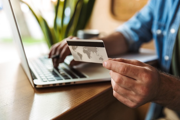 Homem europeu vestindo uma camisa jeans, segurando um cartão de crédito e digitando no laptop enquanto trabalha em um café dentro de casa