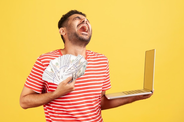 Homem excitado extremamente feliz em camiseta listrada, gritando e rindo segurando laptop e muito dinheiro desfrutando de jackpot vencedora de um estúdio interno isolado em fundo amarelo