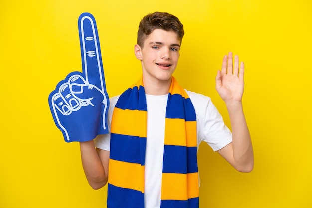 Homem fã de esportes adolescente isolado em fundo amarelo saudando com a mão com expressão feliz