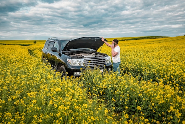 Homem falando ao telefone perto do carro com o capô aberto no campo de colza florescente