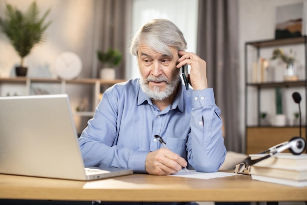 Homem falando no celular enquanto faz anotações na tela do laptop